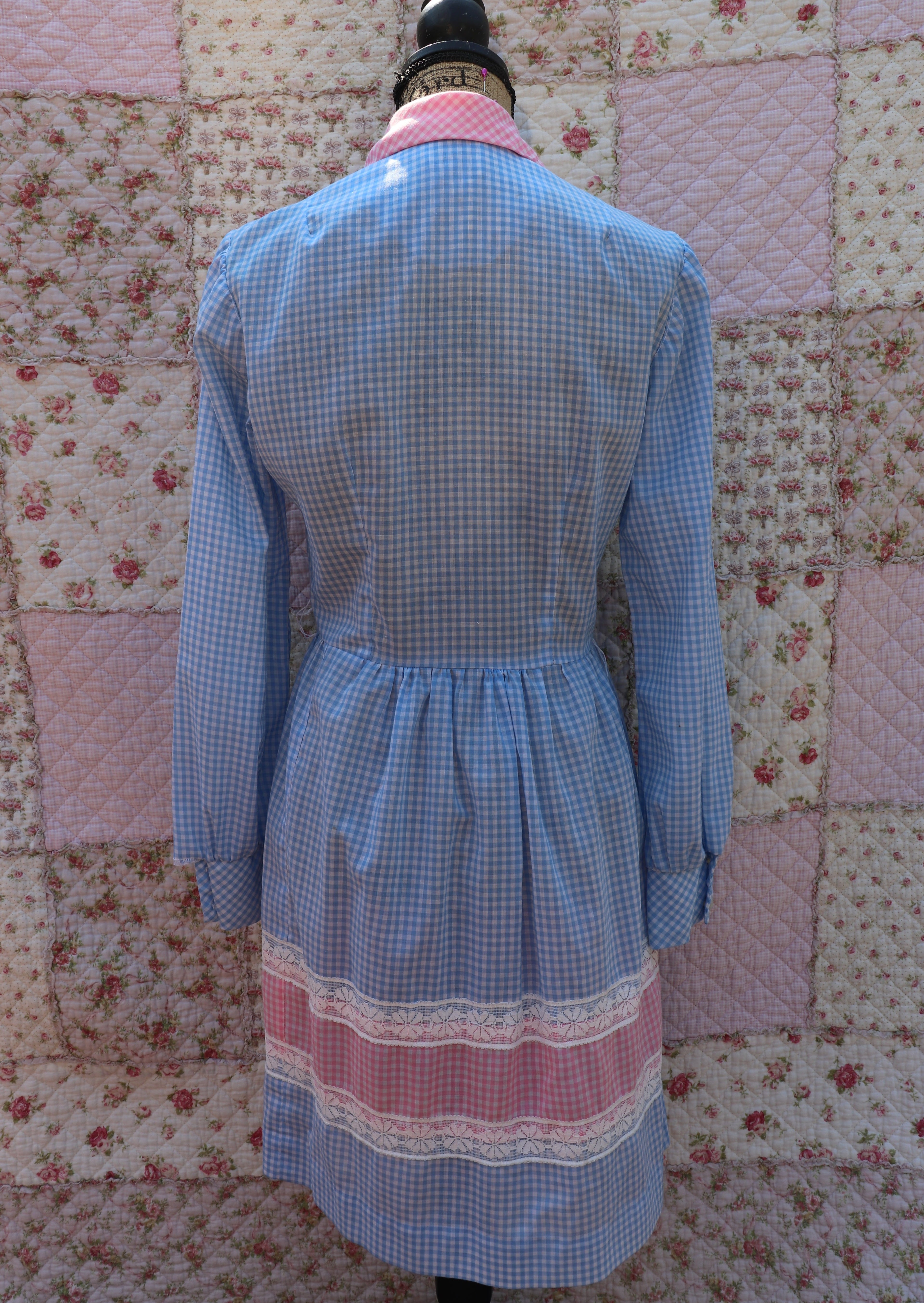 Back view of this dress from J&J Classics! This 1960s dress features a playful blue and white gingham print, accented with pink and white gingham. The long sleeves and collar add a touch of sophistication, while delicate floral lace details add a hint of sweet charm. Get ready to turn heads and spread some flower power with this dress!
