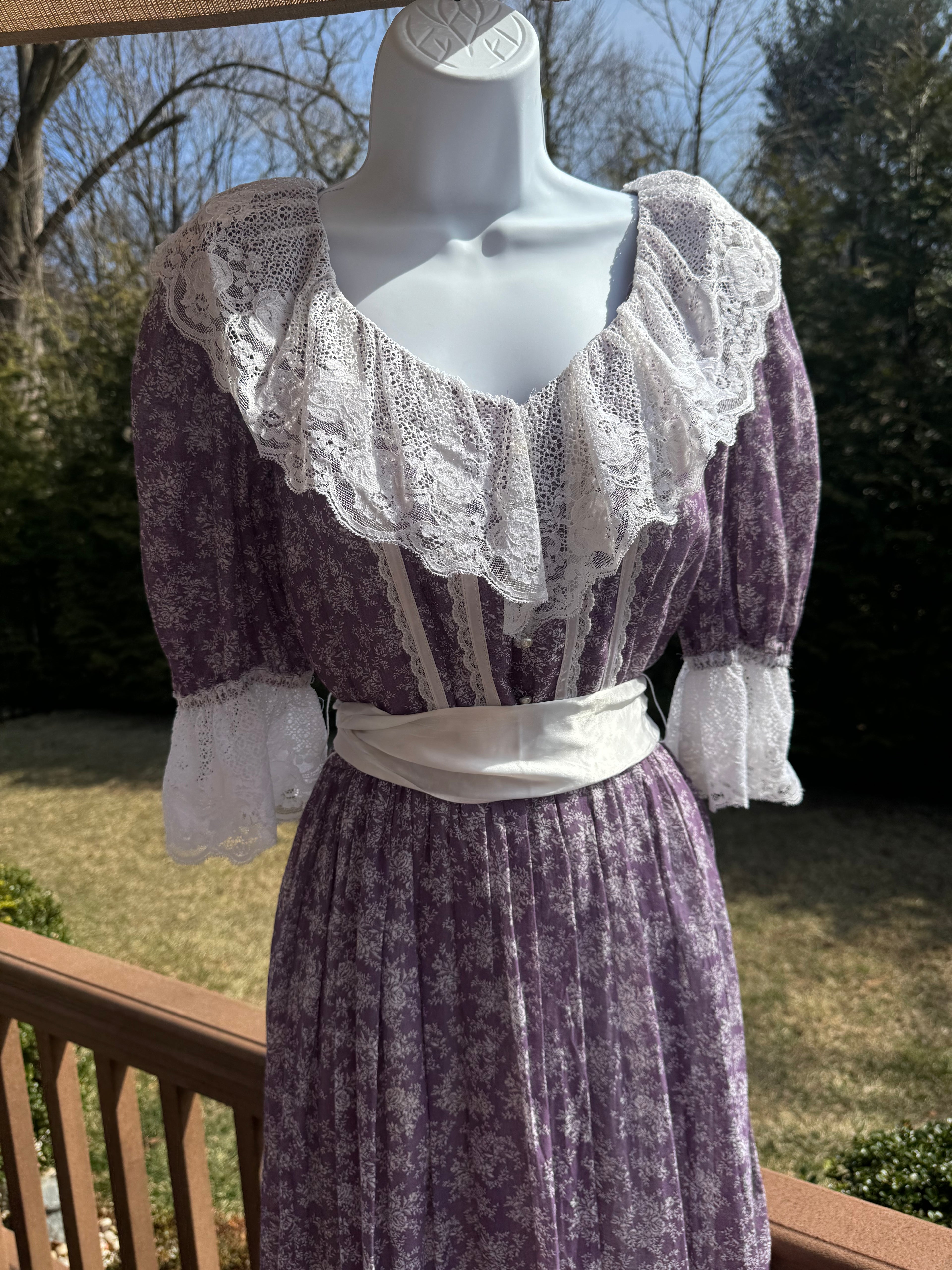 Float like a fairy in the faye dress! This gorgeous purple 1970s Gunne Sax dress is adorned with a dainty white floral pattern and crisp white lace trim. The button front adds an extra touch of unique fun and the attached white ribbon belt adds an extra touch of glamor. Marked as a size 9.