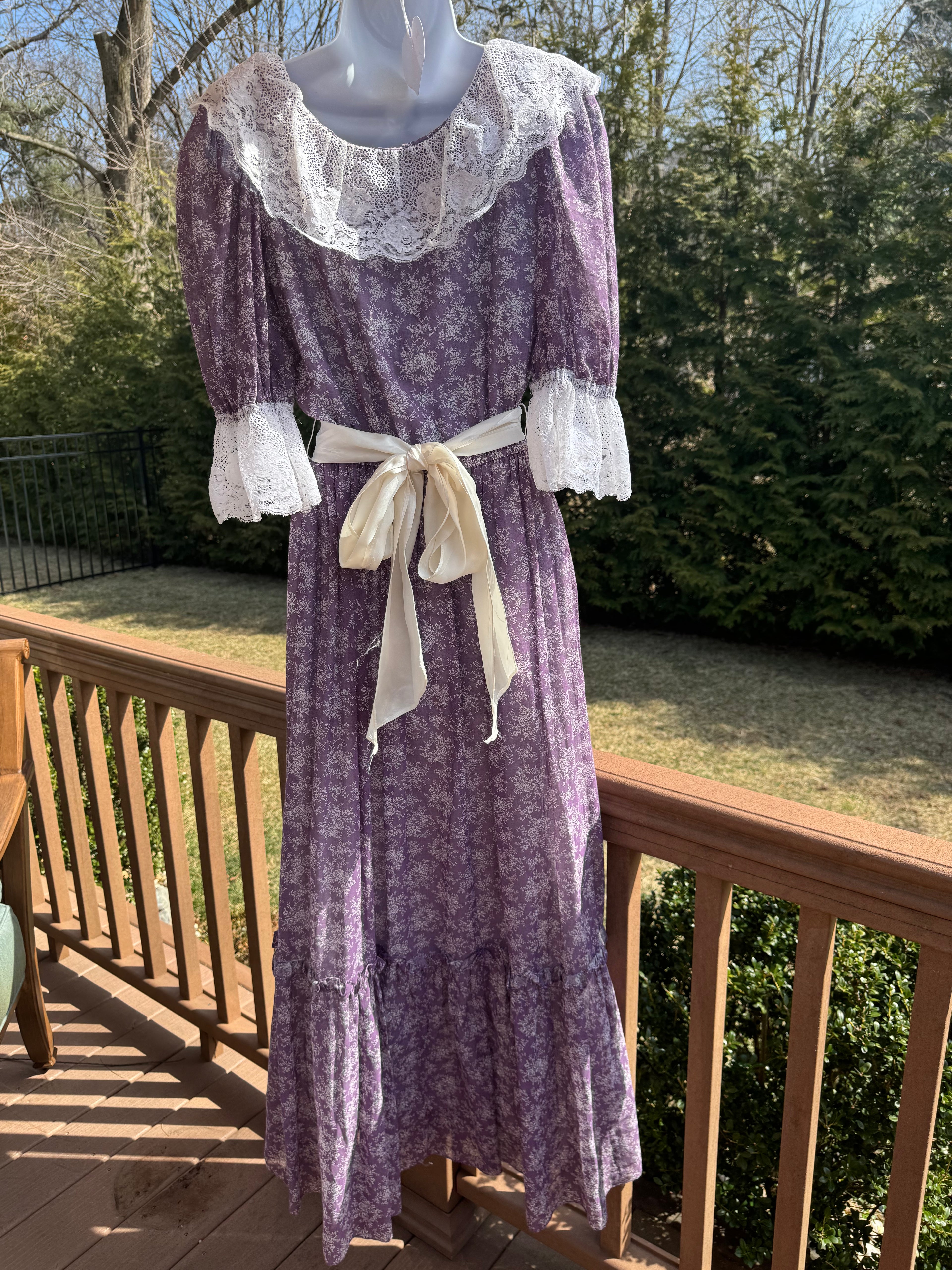 Back view of the faye dress. This gorgeous purple 1970s Gunne Sax dress is adorned with a dainty white floral pattern and crisp white lace trim. The button front adds an extra touch of unique fun and the attached white ribbon belt adds an extra touch of glamor. Marked as a size 9.